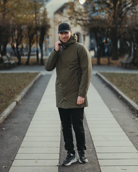 Muž v zelené zimní bundě a černé čepici stojí na chodníku v parku za chladného dne.